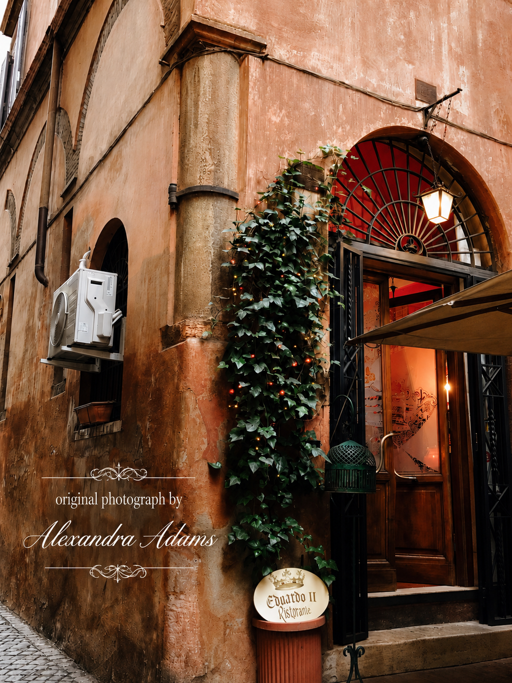 Original photograph by Alexandra Adams of a rustic European restaurant entrance with warm terracotta walls, ivy, arched ironwork and glowing lanterns, featuring an elegant white photography credit overlay