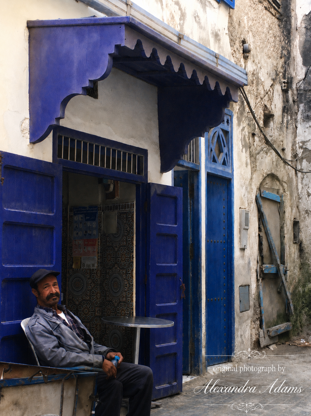 Original photograph by Alexandra Adams showing a seated man outside a vivid cobalt-blue doorway on a weathered Mediterranean street, with an elegant white photography credit positioned at the bottom right.
