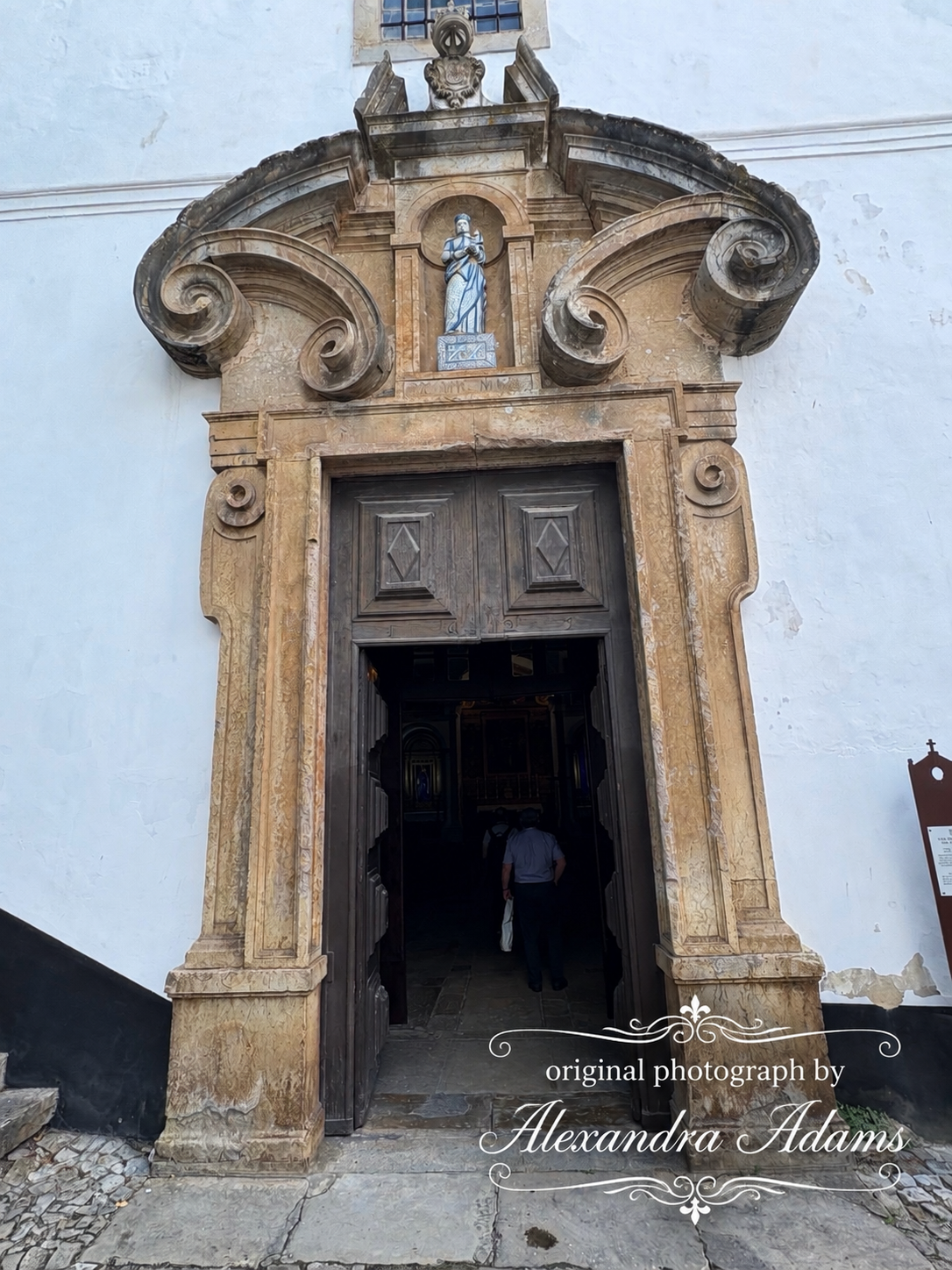 Original photograph by Alexandra Adams showing an ornate historic stone doorway with sculptural scrollwork, a small religious figure above the entrance and open wooden doors leading into a shadowed interior.