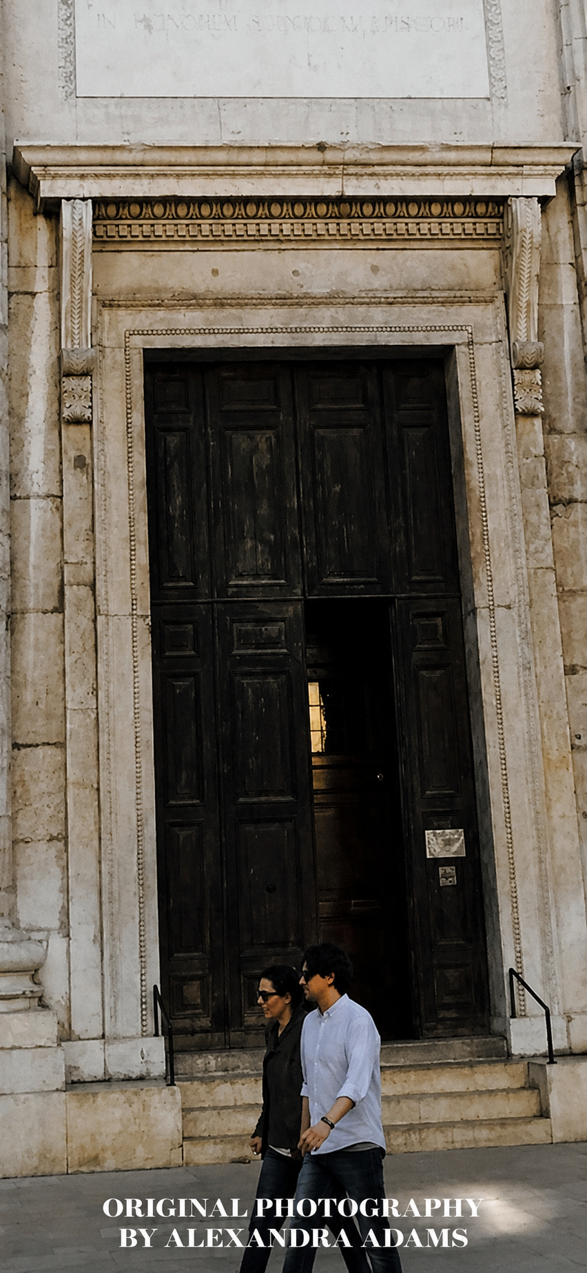 Original photograph by Alexandra Adams showing two people walking past a monumental dark wooden doorway set within an ornate pale stone façade, with bold white photography credit text at the bottom