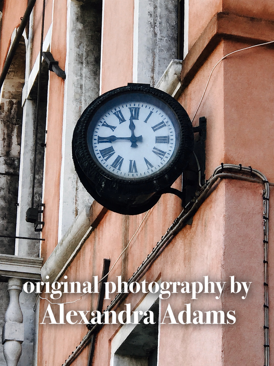 A vintage-style round wall clock with a black metal frame and Roman numerals mounted on a peach-colored building facade. White text at the bottom reads 'original photography by Alexandra Adams'.