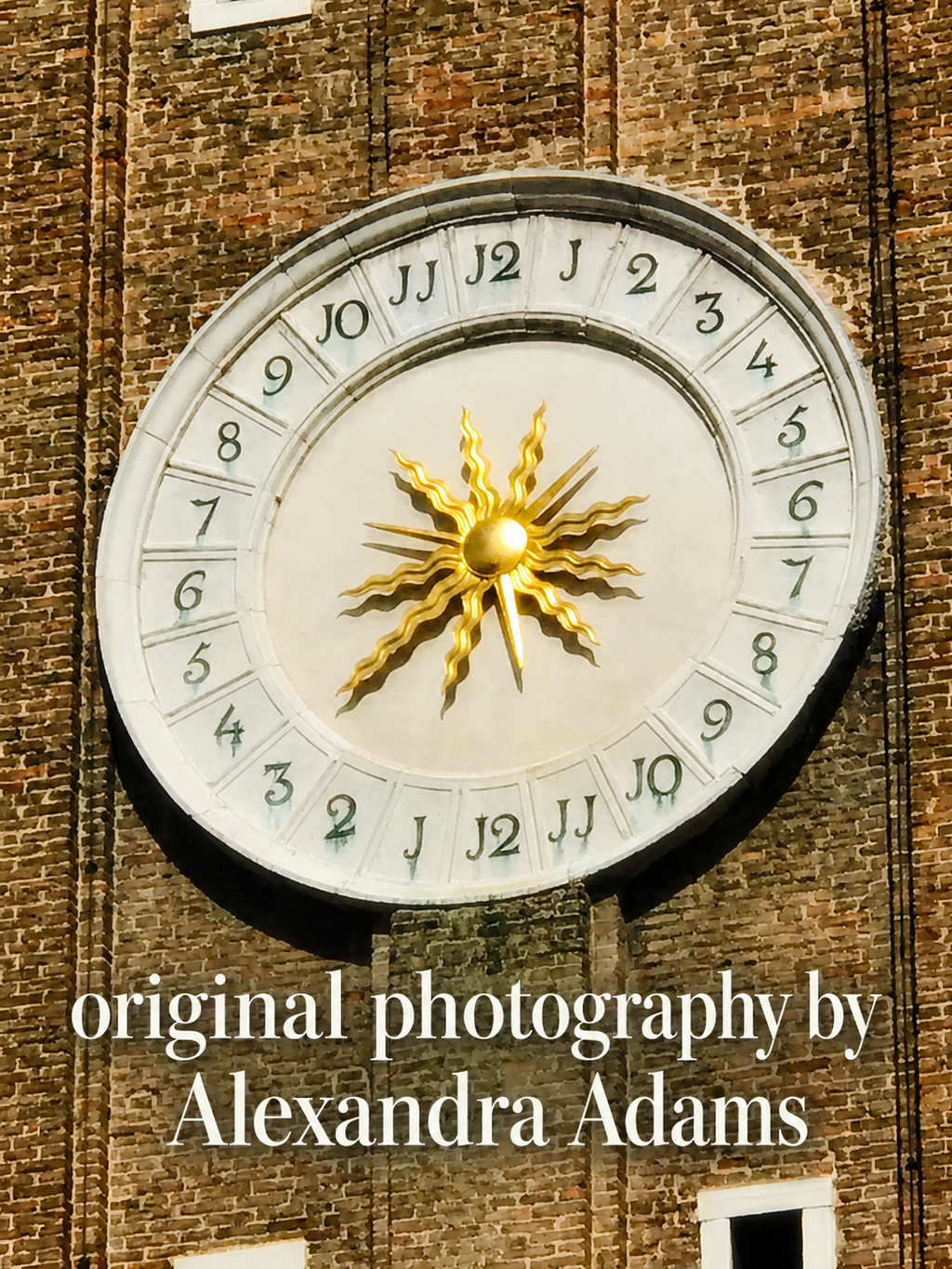 A large circular clock face mounted on a brown brick wall, featuring a gold sunburst center and black Arabic numerals. White text at the bottom reads 'original photography by Alexandra Adams'.