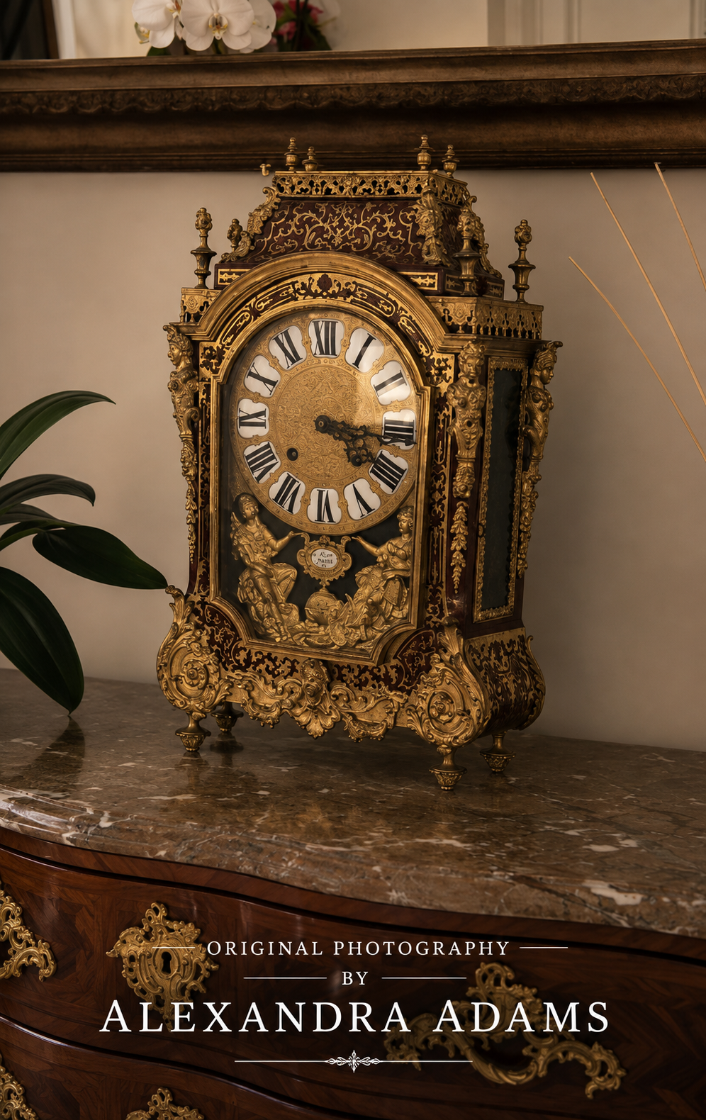 An ornate antique-style gold mantel clock with Roman numerals sits on a polished marble cabinet in an elegant interior setting. The clock features intricate gold filigree detailing and a white clock face.
